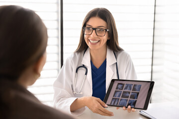 Smiling female doctor providing healthcare services, consulting patient, discussing x-ray results on digital tablet, explaining treatment plan, giving health recommendations in medical clinic office