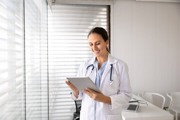 Smiling female doctor working on digital tablet standing in examination room at hospital office. Healthcare assistance services, telemedicine application use, online communication with clinic patients
