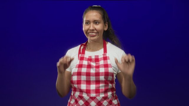 Young woman cook wearing red check apron cupping ear and gesturing with hands against blue studio; curiosity everyday cooking.