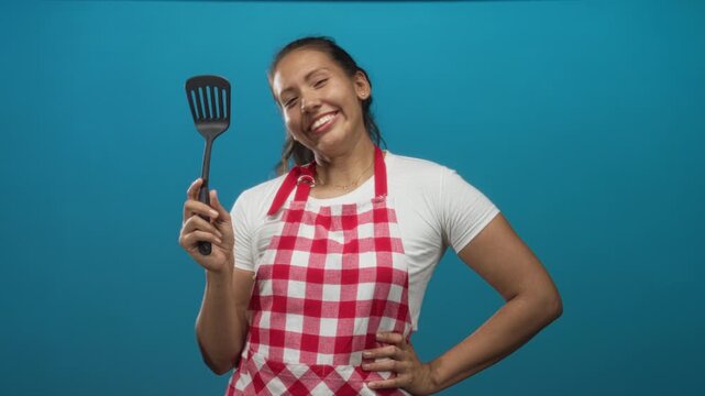 Young hispanic woman cook holding spatula and smiling in blue studio, wearing red check apron and white t shirt; cheerful cooking.