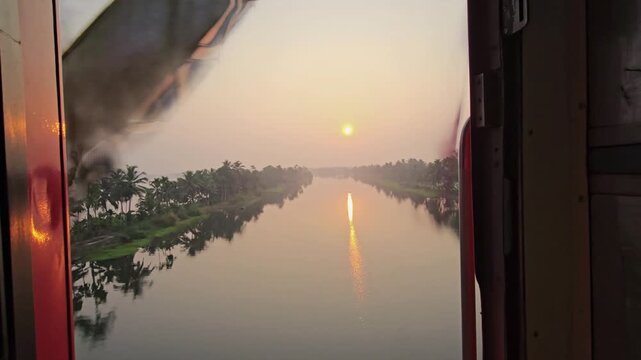 A serene sunset over Kerala's backwaters, viewed from a train. Golden light reflects on tranquil waters lined with lush palms near Alleppey, as a metal truss bridge passes by the window. Captivating.