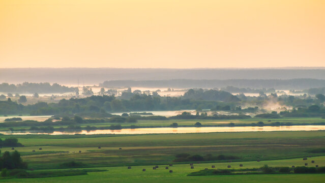 Green fields and meadows with trees and river at pink foggy sunrise. Beautiful summer landscape.