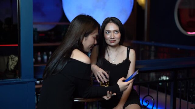 Asian woman friends talking and meeting at a bar. Two young ladies looking at smartphone and laughing in a nightclub. Social media, nightlife, and friendship.