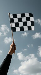 Fototapeta premium Person holding checkered race flag high against a blue sky for motorsport victory and achievement concept, low angle shot of hand with winning flag outdoors