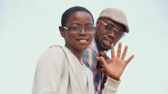 Black father and son smiling and waving on bright outdoor sky background, casual clothing, son wearing glasses, father wearing cap, playful hand wave, genuine joy, close bond, family travel mood, warm