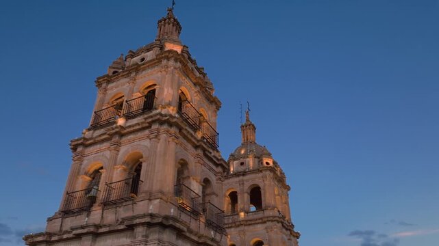 Cathedral Basilica of the Immaculate Conception in Durango, Mexico during golden sunset. Twin bell towers with ornate baroque architecture illuminated against deep blue twilight sky with wispy clouds.