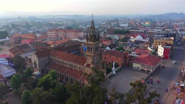 Aerial view of historic church with tall spire. Red rooftops surround the church, streets below. Cityscape stretches into hazy hills. Peaceful morning light illuminates architecture