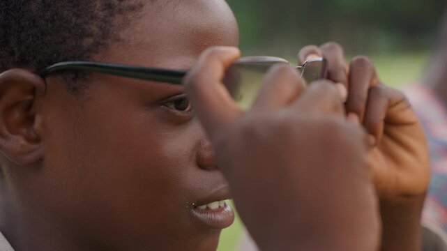 Closeup black son adjusting sunglasses outside, young student tries on dark lenses while father watches from blurred background, warm afternoon light, park grass and gentle smile convey tentative
