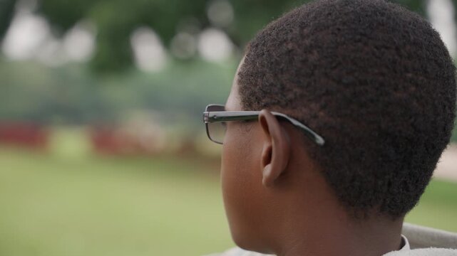 Closeup black son wearing glasses looking over shoulder, soft bokeh park background, short afro haircut, calm afternoon light, candid profile shot conveying introspection, gentle texture of skin,