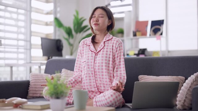 Asian adult woman practices mindfulness meditation sitting lotus pose in pink pajamas on sofa at home for peace wellness relaxation