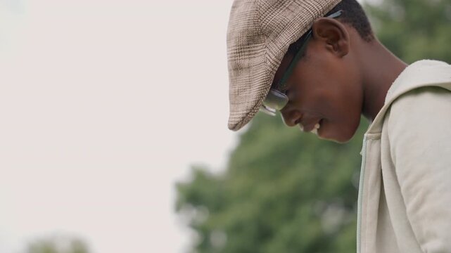 Black son in park looking down, wearing cap and hoodie candid profile shot with soft trees background and gentle daylight introspective mood, nostalgic atmosphere, subtle smile and thoughtful