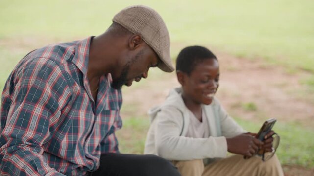 Black father and son with phone smiling and laughing while sharing mobile game, father acting as playful coach, casual cap and check shirt, grassy park setting, joyful candid interaction