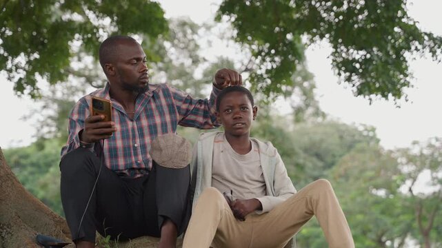 Black father and son laughing on tree branch, father ruffles sons hair while they sit under oak, playful teaching and tender mentorship, natural light, casual clothes, carefree childhood scene
