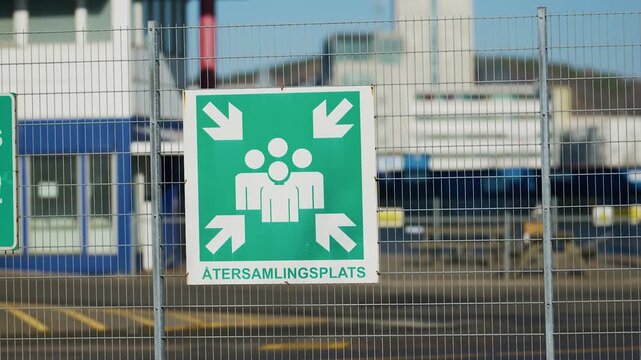 Emergency assembly point safety sign on metal fence indicating evacuation meeting area outdoors