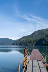 Obraz premium Wooden dock extending into calm reflective waters with forested mountains and blue sky at Lake Crescent in Olympic National Park, Washington State, USA. Scenic Pacific Northwest landscape