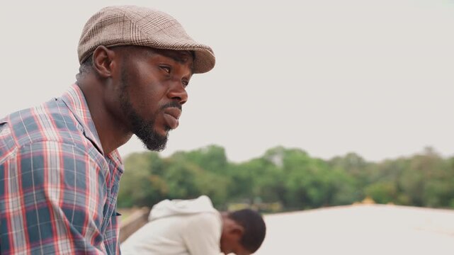 Black father and son fishing lakeside, plaid shirt dad wearing cap leans over boat while son watches, green treeline backdrop, calm overcast sky, gentle water reflections, intimate teaching moment,