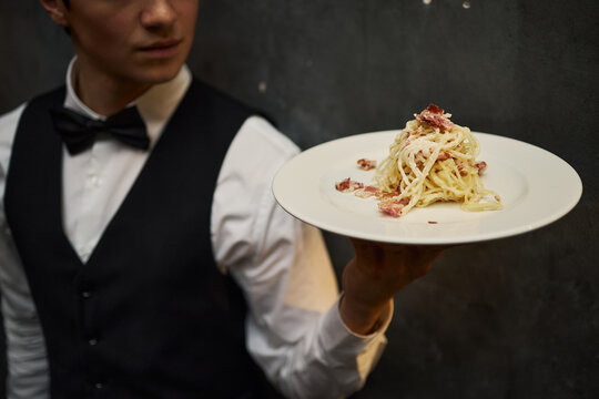 Young adult Caucasian waiter presenting plated pasta dish in restaurant service setting, highlighting hospitality and fine dining. Useful for menu design, catering ads, food service marketing