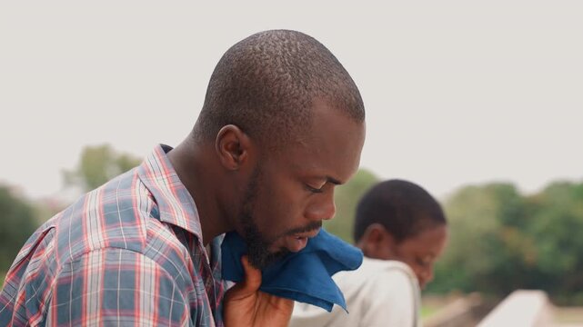 Father and son outdoors wiping sweat, father dabs forehead with blue towel while son smiles in background rooftop setting warm light plaid shirt candid closeup emotional moment of care and relief