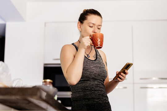 Woman in grey sportswear drinking coffee and using smartphone indoors