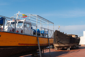 Fototapeta premium Bright yellow passenger ferry boat with metal cage seating structure rests beside a historic wooden black vessel upon a dry dock platform under a clear blue sky in summer daylight.