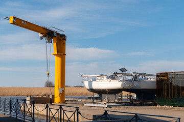 A vibrant yellow gantry crane stands prominently at a boatyard, ready for lifting operations, with several white sailboats drydocked on stands for seasonal maintenance or storage under a clear sky. © Indre