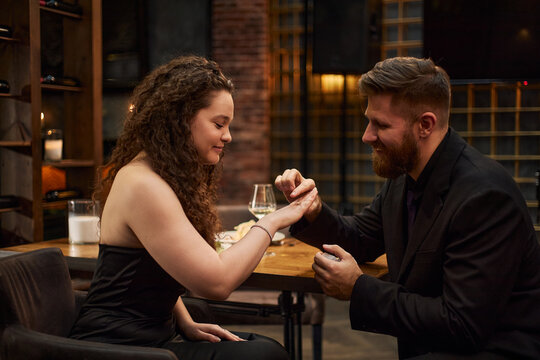 Caucasian young adult man proposing to Caucasian young adult woman, placing engagement ring on finger during restaurant date, symbolizing commitment. Ideal for wedding marketing