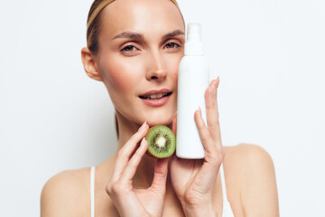 Woman with natural makeup holds cotton pad and kiwi slice near face, skincare routine concept, she touches cheeks, bright white background, beauty wellness self care fresh glow expression