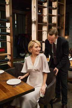 Vertical shot of middle aged Caucasian couple arriving for fine dining, man assisting woman with seating at restaurant table. Suitable for luxury hospitality, dating, celebration marketing