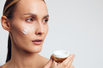 Young woman applying cream on face holding jar, skincare routine, natural beauty, clean skin, soft lighting, neutral background, focused expression, healthy lifestyle