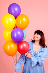 Obraz premium Happy young brunette woman in a blue dress with colorful balloons on a pink background
