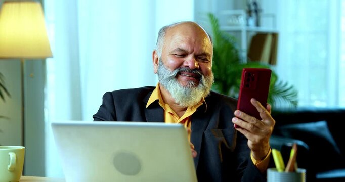 Indian senior businessman or corporate executive engaging in a friendly chat on his smartphone while simultaneously working on a laptop at his desk in a cozy office with moody lighting.
