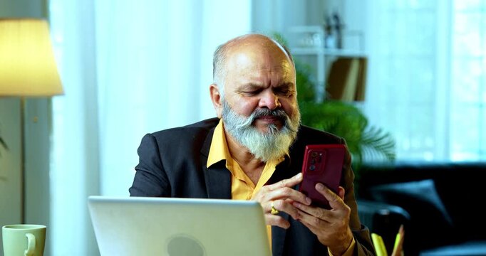 Indian senior businessman or corporate executive engaging in a friendly chat on his smartphone while simultaneously working on a laptop at his desk in a cozy office with moody lighting.