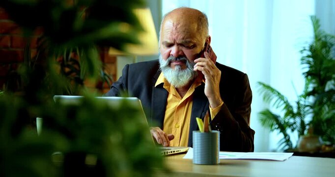 Indian senior businessman or corporate executive multitasking by speaking on a smartphone while actively typing on a laptop at his desk in a cozy office with professional moody lighting.