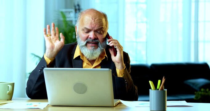 Indian senior businessman or corporate executive multitasking by speaking on a smartphone while actively typing on a laptop at his desk in a cozy office with professional moody lighting.