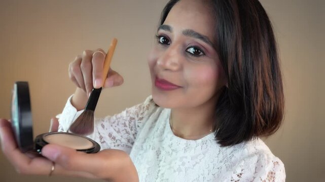 Close up of happy beautiful young Indian woman applying make up hold powder compact and brush look at camera smiling, isolated over grey studio background.