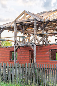 Unfinished red brick house features wooden structural frame for its second story and roof. Weathered wooden picket fence stands in front of the building