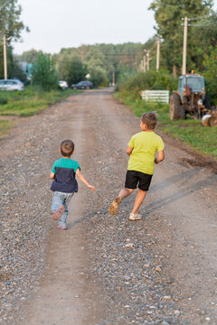 Two boys running on gravel road racing toward distant farmhouse under warm sunlight, siblings laughing, kicking up dust, tractor parked beside lane, lush