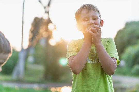 Boy blowing on hands in golden light, backlit portrait with sun flare, gentle expression and soft bokeh, warm evening mood in neighbourhood park setting