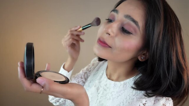 Close up of happy beautiful young Indian woman applying compact powder with brush to beautiful face, isolated over grey studio background.