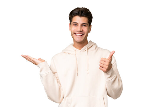 Young caucasian handsome man over isolated background holding copyspace imaginary on the palm to insert an ad and with thumbs up