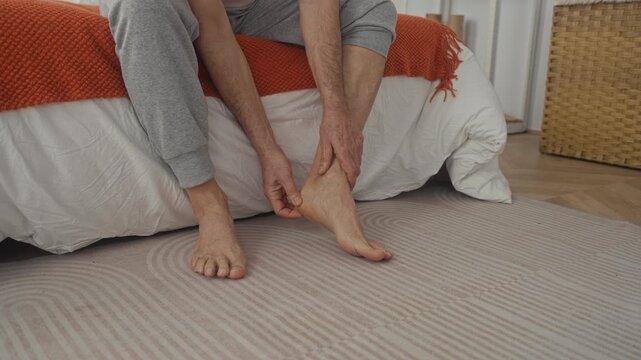 Man massaging foot on bed in bedroom with orange blanket and woven basket, wearing grey sweatpants barefoot; comfort relief.
