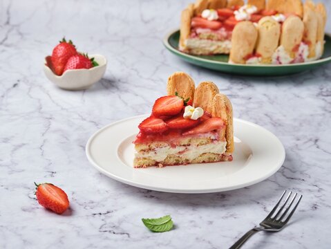 Round tiramisu cake with fresh strawberries and mascarpone cream decorated with marshmello flowers on a green plate on a light marble background. No-bake desserts.