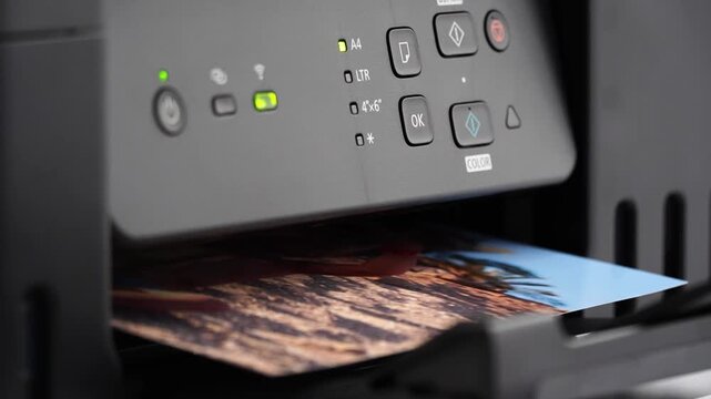 Printer outputs vibrant video with warm sunset tones. Paper emerges slowly from printer's exit tray. Control panel glows with green status lights. Print job completes with crisp, detailed image