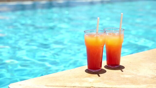 Two colorful drinks sit beside a bright blue pool. Sunlight glints on the water's surface. Crisp shadows fall on the poolside stone. Tropical vibes radiate from the scene. Perfect for vacation