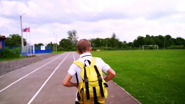 Child walks away on running track, arms open wide. Bright yellow backpack contrasts with green grass and blue buildings. Cloudy sky adds soft, calm atmosphere to the scene. Child appears free, joyful