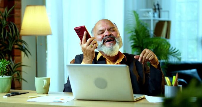 Indian senior businessman or corporate executive multitasking by speaking on speakerphone via his smartphone while actively typing on a laptop at his desk in a cozy office with moody lighting.