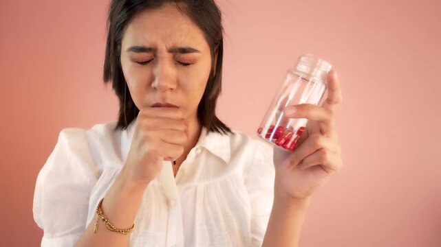 Close up of ill sick young Asian woman coughing sore throat, suffering cold or flu virus holding bottle look at capsules hesitation hate, dislike medicine, isolated against grey studio background.