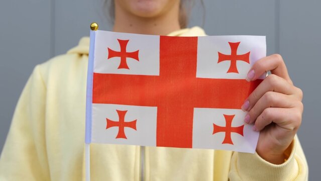 Young Caucasian cute girl is keeping flag of Georgia in her hands and waving and smiling at camera. Zoom out, close up, grey background