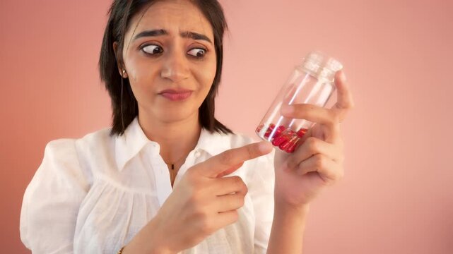 Close up of unhappy young Asian woman holding bottle look at capsules hesitation or hate, dislike medicine, isolated against grey studio background.
