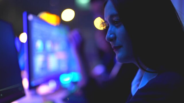 Focused female specialist with glasses scrutinizing complex coding and AI algorithms on screen while professional colleagues work in the background at a high tech night office hub.